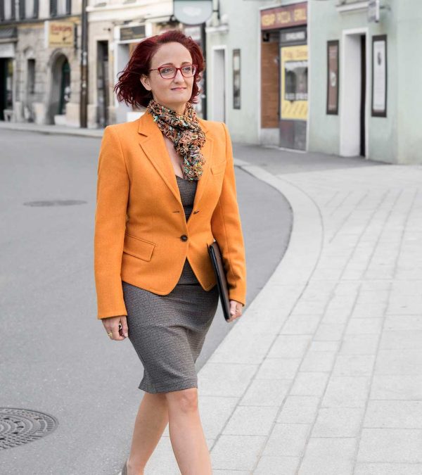 Dame im Business-Look mit sommerlichen Loop-Schal passend zum organgen Blazer und dem braun-grauen Rock in der Fußgängerzone einer Stadt