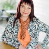 glückliche Frau im Freizeitlook mit orangem Loop, orangem Shirt und floralbedruckter Jacke in Zartgrün, Schwarz und Orange
