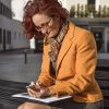 Geschäftsfrau mit buntem Sommerloop von il-design passend zum orangen Blazer und dem grauen Rock auf einer Bank sitzend ins Handy tippend